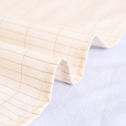 A close-up of a bejge-colored fabric with a silver fibre, thin grid pattern, folded over to show its texture and stitching, resting on a light white surface.