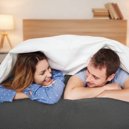 A smiling woman in a blue star-patterned shirt and a man in a light blue t-shirt lying side-by-side on a dark gray perforated mattress topper grounding sheet under white sheets.